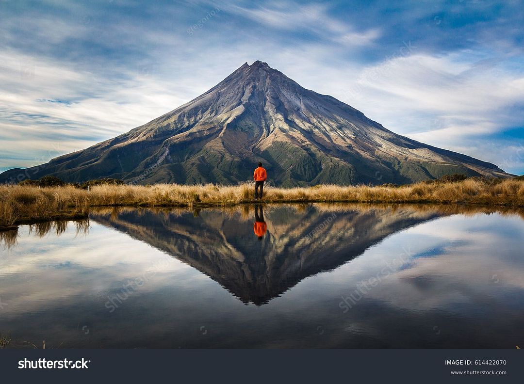 stock-photo-a-mountaineer-with-an-orange-dress-in-a-symmetric-picture-looking-to-the-taranaki-volcano-in-the-614422070