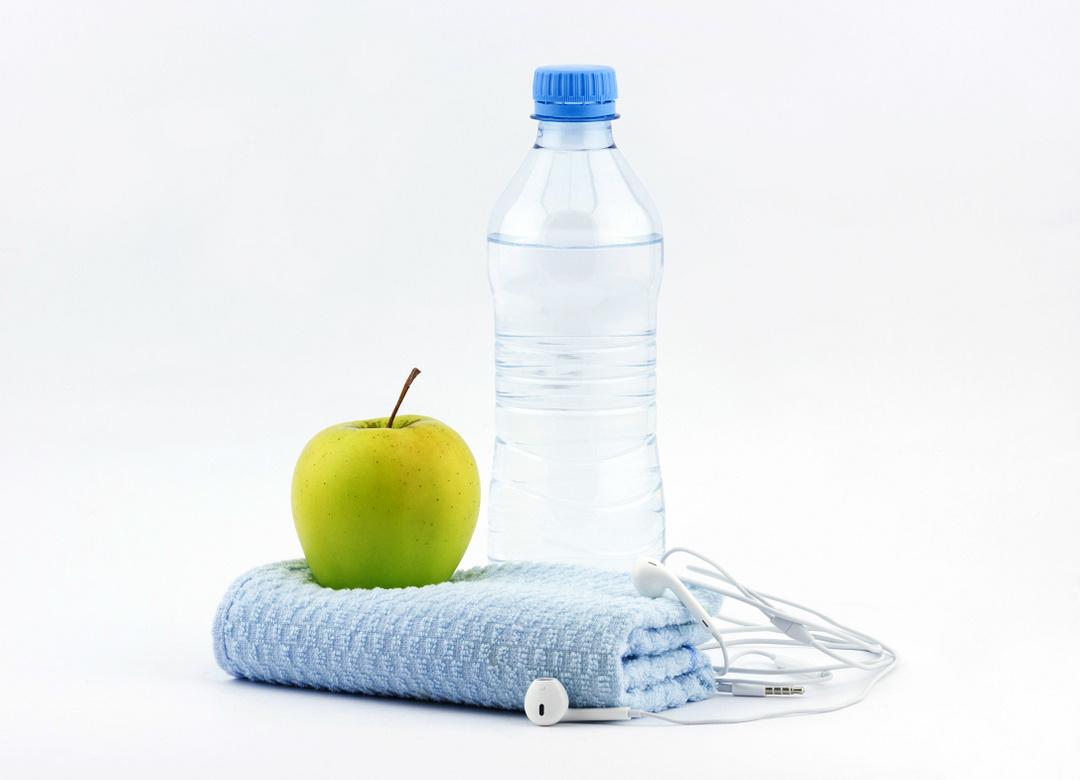 Healthy lifestyle concept. Bottle of water with green apple, fitness towel and headphones on white background