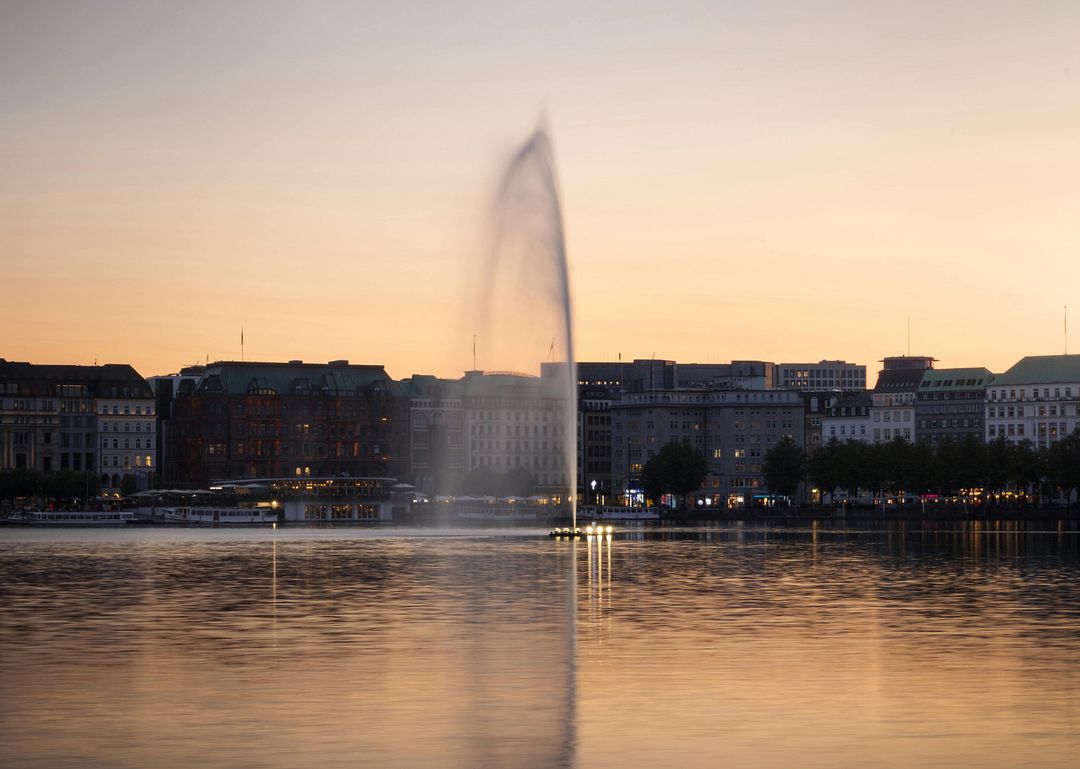 The image shows the Geneva Fountain in Geneva gushing out of the water against a sunset background.  The Geneva Fountain in Geneva (also called Do) is an important landmark of the city and one of the largest fountains in the world.   History: the fountain was originally built at the end of the XIX century as an additional structure to the hydraulic factory, a source of energy for the city's industries. His task was to dump excess water into the lake, which accumulates on weekends and in the evening in the hydraulic system.   Specifications:  The height is 147 meters.  The flow rate is 500 liters per second.  The power consumption of each of the two pumps is 500 kW.  The jet speed is 200 km/h.  The fountain is illuminated by 12 floodlights, the lighting power is 9,000 watts.  Features:  Under the influence of wind, combined with sunlight, the water column changes shape and colors.  The type of jet can take the form of a column, or it can crumble into splashes, fanning almost to the middle of the lake.  Working hours: since 2003, the fountain has been operating daily according to a special schedule: during the winter months during the daytime, in spring, autumn and summer until sunset on weekdays and almost until midnight on weekends and holidays, in November the fountain is completely turned off. It is also turned off in very strong winds and frosts.