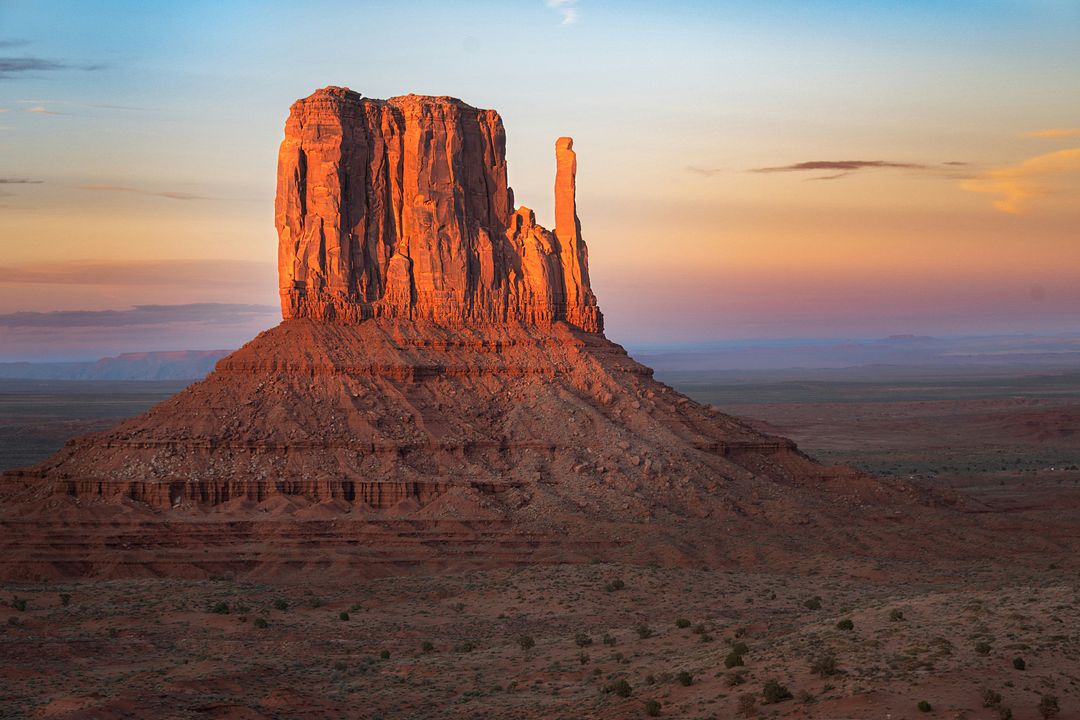 The warm glow of the setting sun accentuates the majestic silhouette of West Mitten Butte in Monument Valley, casting a tranquil scene over the vast Arizona landscape.