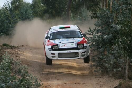 The image shows a white Toyota Celica GT-Four car with logos on the hood, moving along a dirt road.  Toyota Celica GT-Four is a Celica model in a liftback body, with a turbocharged 3S—GTE engine and a permanent all-wheel drive system.   The car was created to participate in the World Rally Championship. 13 The rules of this championship required the manufacturer to produce road versions of the car in sufficient quantities.