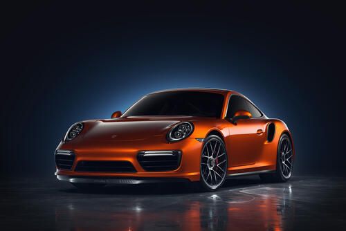 The image shows an orange Porsche 911 Turbo S Exclusive Series sports car with a two-door body.  The Porsche 911 Turbo S Exclusive Series is the most powerful and rare variant of the 911 Turbo S. Its release was limited to 500 copies.   The Exclusive Series differs from the basic version of the model in the following:  The engine power has been increased by 27 horsepower. The 3.8-liter six-cylinder turbo engine develops 607 horsepower and 750 Nm of torque.  Acceleration from a standstill to 100 km/h in 2.9 seconds, and to 200 km/ h in 9.6 seconds. The maximum speed of the sports car is 330 km/h.  The equipment includes 20-inch center-mounted wheels, ceramic brakes, PASM adaptive suspension, full-control chassis and Sport Chrono package.  Other features of the model:  The body color is unique, golden. Accents of the same color are also present in the interior.  In the cabin there are carbon fiber inserts with copper addition, double—layer perforated leather on sports chairs with electric adjustments in 18 directions.  Each car was accompanied by a watch from Porsche Design, in the manufacture of which materials such as titanium and carbon were used