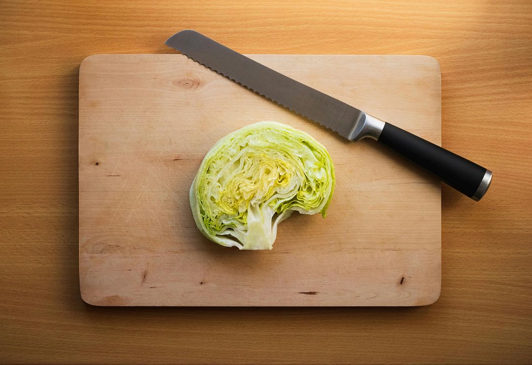 The image shows a head of white cabbage with a knife on a chopping board.  To cut white cabbage on a chopping board with a knife, you can follow these recommendations:  Prepare the vegetable. Rinse with running water and rub with your hands to remove impurities and pesticides. Wipe the washed head with a paper towel. Remove damaged leaves that have spots, holes and discolored ones.  Choose a knife. A stainless steel tool is suitable and sharp enough. It is not recommended to use a carbon metal knife, as it can leave black spots on the cut.  Cut the head into quarters. Press the vegetable tightly against the board and hold it with the fingers of one hand. Make an incision in the center in one smooth motion.  Remove the core.  Hold the quarter of the head vertically, with the bottom side with the stalk down. Cut the stem diagonally with one firm incision. At the same time, you do not need to dip the knife deep into the leaves.  Chop or chop the vegetable.  Place the cut down on a cutting board. 3 Bend your fingers and hold the cabbage with them so that their joints, and not the tips of your fingers, are located closer to the blade of the knife.  Chop the cabbage, starting from the outside and gradually moving towards the middle.  Cut the pieces. To keep the pieces short, cut the quarter across. To cut into long strips, cut lengthwise